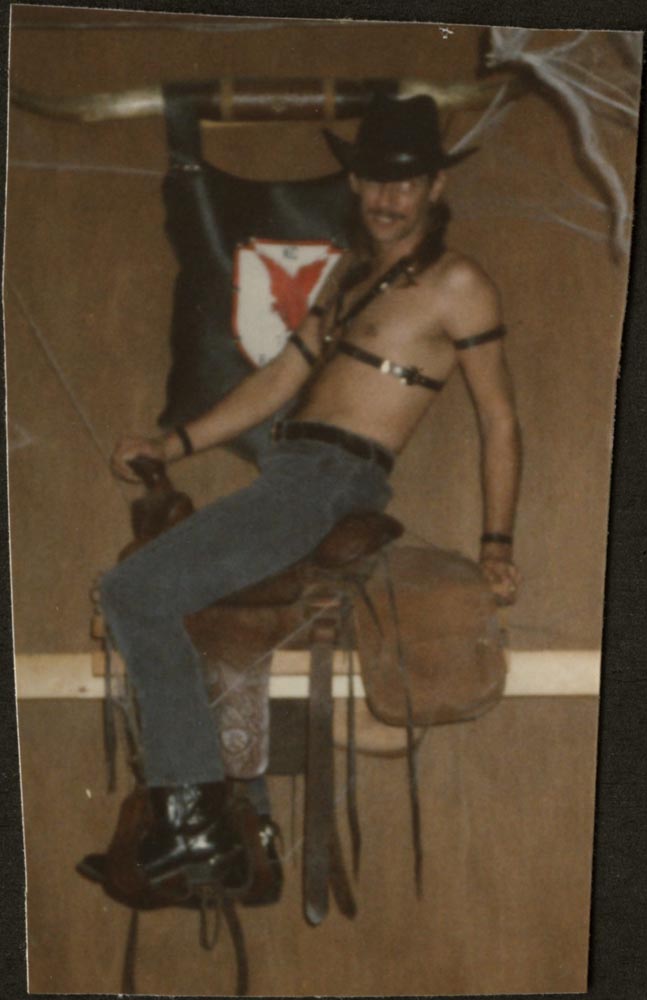 Shirtless person in bondage gear riding a horse saddle strapped to a bar stool